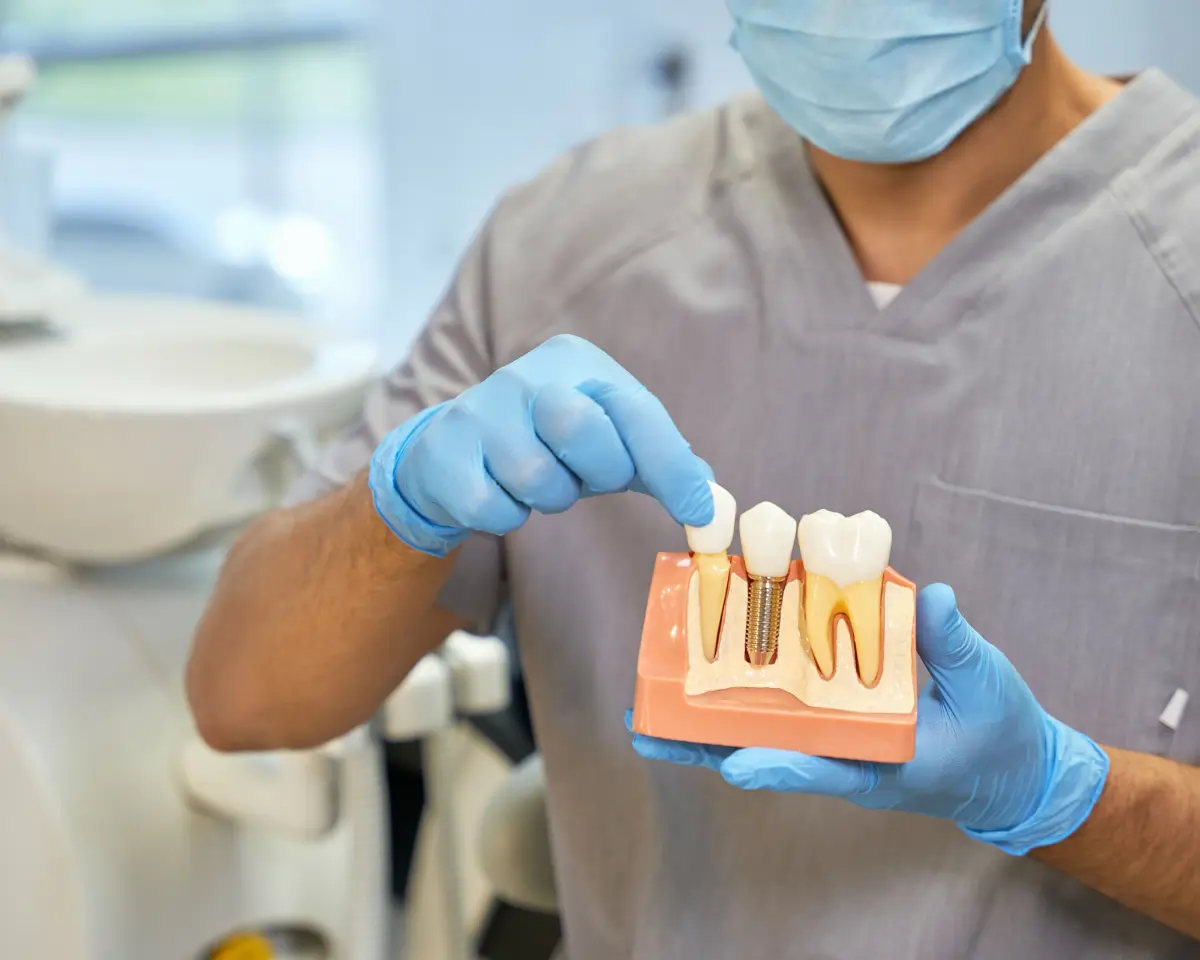 Gloved dental professional in gray scrubs holding a cross-section implant model and pointing at the crown component in a clinical setting